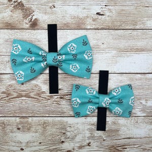 May include: Two turquoise bow ties with a white floral pattern. The bow ties are attached to black elastic bands.