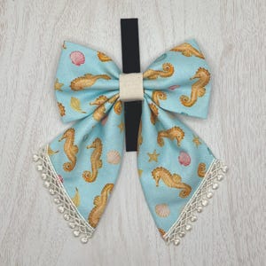 May include: A large, light blue bow with a seahorse and seashell pattern. The bow has a black band and a beige center. White lace trim adorns the bottom edges. The bow is likely for a child's hair.