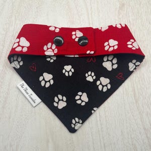 May include: A dog bandana with a red and black paw print design. The top section is red with white paw prints and black snaps. The bottom section is black with white paw prints and small red hearts. A white tag reads "The Three Tuxedos."