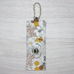 Könnte beinhalten: Ein weißer Stoff-Lesezeichen mit einem Bienen-, Waben- und Blumendesign. Das Lesezeichen hat einen schwarzen und gelben Quasten.