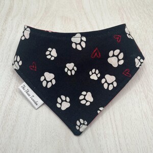 May include: A black dog bandana with white paw prints and small red hearts scattered across the fabric. The bandana has a triangular shape and a tag that reads "The Three Tuxedos".