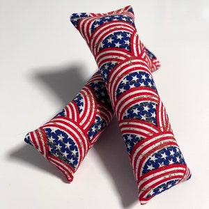 Puede incluir: Dos juguetes para gatos de tela roja, blanca y azul con un patrón de estrellas. Los juguetes tienen forma de cilindros largos.