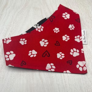 May include: A reversible dog bandana with a red side featuring white paw prints and black hearts. The reverse side is black with white paw prints. The tag reads "The Three Tuxedos".