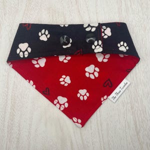 May include: A reversible dog bandana with a black top section and a red bottom section. The black section has white paw prints. The red section has white paw prints and black hearts. Two black buttons secure the bandana.