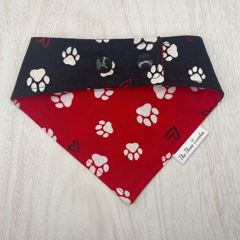May include: A reversible dog bandana with a black top section and a red bottom section. The black section has white paw prints. The red section has white paw prints and black hearts. Two black buttons secure the bandana.