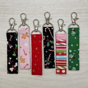 May include: Six Christmas-themed keychains with different patterns. The keychains are made of fabric and have silver metal rings. The patterns include candy canes, gingerbread men, snowflakes, stars, and holly.
