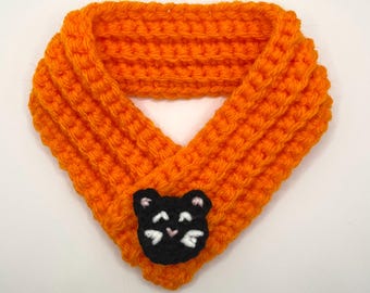 Peut inclure: Écharpe pour animaux de compagnie en crochet orange vif avec un visage de chat noir. L'écharpe est faite d'un fil épais et texturé, conçue pour s'adapter au cou d'un animal de compagnie. Le visage du chat a des détails blancs et des accents roses.