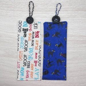 May include: Two fabric bookmarks with black buttons and black string ties. One bookmark is white with black and orange text that says "w wow DU GOOD WOOF N WOW CATCH JUMP Sniff BO WOOF WOOF Dog BEG GROWL ARF ARF SPEAK goo BOW WOW PLAY roll over PLAY BEG LUCK GOOD DOC". The other bookmark is blue with black cats printed on it.