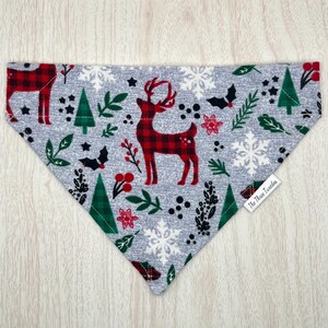 Könnte beinhalten: Ein graues Hunde-Bandana mit festlichem Muster. Es zeigt rot karierte Hirsche, grüne Bäume, Schneeflocken und florale Akzente. Das Bandana hat ein Etikett mit der Aufschrift "The Three Tweeds".