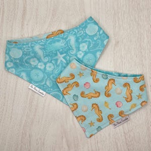 May include: Two blue pet bandanas with sea-themed patterns. One features seahorses, shells, and starfish, while the other has a similar design with seahorses, shells, and a crab. Both bandanas have a tag that reads "The Three Tunas".