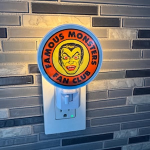 May include: A round night light with the words "FAMOUS MONSTERS FAN CLUB" encircling a yellow and red monster face. The night light is plugged into a white electrical outlet on a gray and black tiled wall.
