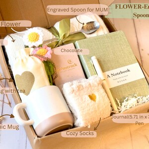 May include: A brown gift box filled with a variety of items, including a ceramic mug, cozy socks, a journal, a pen, a burlap bag with tea, chocolate, and a flower. The box is labeled "Engraved Spoon for MUM" and "FLOWER-Engraved Spoon".