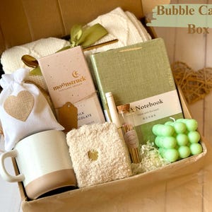 May include: A brown gift box filled with a variety of items, including a white mug, a green notebook, a small bag of chocolate, a white blanket, a green bubble candle, and a small bag of bath salts. The box is labeled "Bubble Candle Box".
