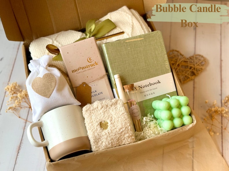 May include: A brown gift box filled with a variety of items, including a white mug, a green notebook, a small bag of chocolate, a white blanket, a green bubble candle, and a small bag of bath salts. The box is labeled "Bubble Candle Box".