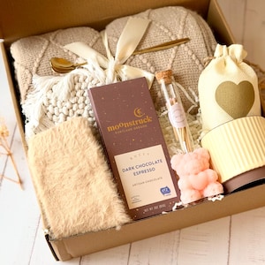 May include: A gift box filled with cozy items, including a blanket, a mug, a bar of dark chocolate espresso, a small bag of matches, and a pink bear-shaped candle. The box is made of brown cardboard and has a white background.