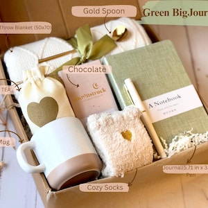 May include: A gift box filled with cozy items, including a knit throw blanket, a ceramic mug, a burlap bag, a bar of chocolate, a journal with a pen, cozy socks, and a gold spoon. The box is decorated with a green ribbon and a gold heart.