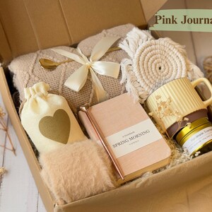 May include: A gift box filled with items in a neutral color palette. The box contains a beige blanket, a small white bag with a gold heart, a journal with the text "A Notebook Spring Morning" on the cover, a gold pen, a small white coaster, a yellow mug, and a small gold candle.