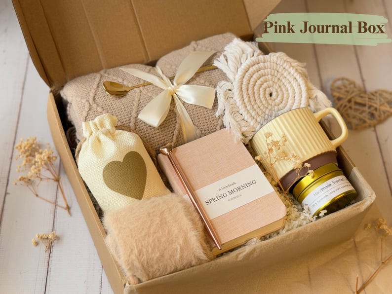 May include: A gift box filled with items in a neutral color palette. The box contains a beige blanket, a small white bag with a gold heart, a journal with the text "A Notebook Spring Morning" on the cover, a gold pen, a small white coaster, a yellow mug, and a small gold candle.