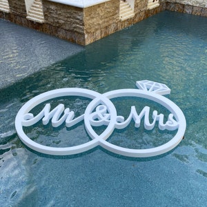 May include: White wooden ring sign with the words "Mr. & Mrs." and a diamond in the center, floating in a blue pool.