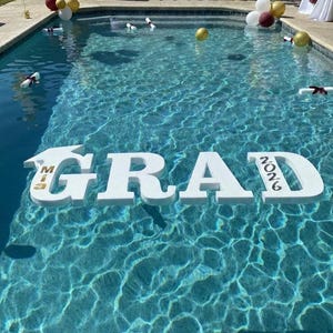 May include: A pool is decorated for a graduation celebration. Large white letters spell out "GRAD" with "MIA" and "2026" in gold. Graduation caps, balloons, and rolled diplomas float in the water.