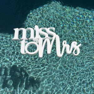 May include: White wooden sign with the words "miss to mrs" and a diamond ring symbol. The sign is floating in a blue swimming pool.