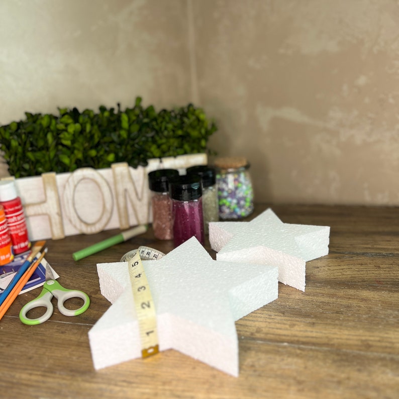 May include: Two white foam star shapes, one on top of the other, with a measuring tape wrapped around the bottom star. The stars are on a wooden surface with craft supplies in the background, including paint, glitter, and a small wooden box with the word "HOME" on it.
