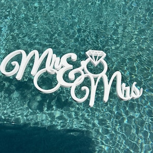 May include: White wooden sign with the words "Mrs & Mrs" and a diamond ring symbol in the center. The sign is floating in a blue swimming pool.