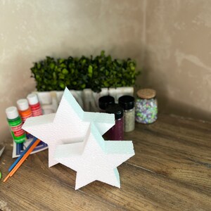 May include: Two white foam star shapes, one on top of the other, on a wooden surface. There are paint bottles and other craft supplies in the background.