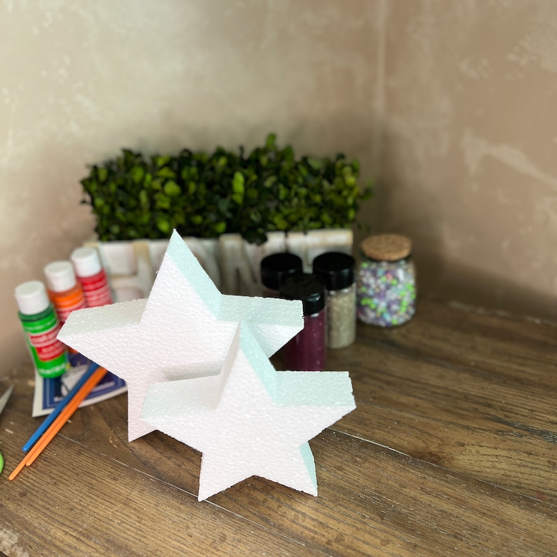 May include: Two white foam star shapes, one on top of the other, on a wooden surface. There are paint bottles and other craft supplies in the background.