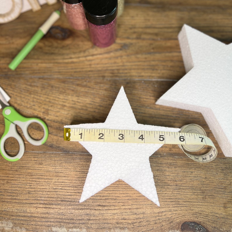 May include: Two white foam star shapes, one larger than the other, are being measured with a tape measure. The tape measure reads 7 inches.