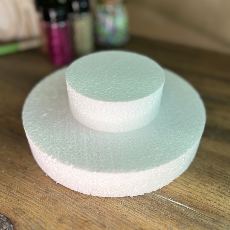 May include: Two white round foam cake dummies stacked on top of each other. The larger dummy is on the bottom and the smaller dummy is on top.