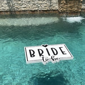 May include: White sign with black text that says "Bride to be" floating in a blue swimming pool.