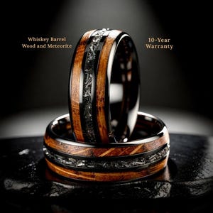 May include: Two rings with a whiskey barrel wood and meteorite design. The rings have a black band with a wood grain pattern and a center of dark gray meteorite. The text "Whiskey Barrel Wood and Meteorite" and "10-Year Warranty" are visible.