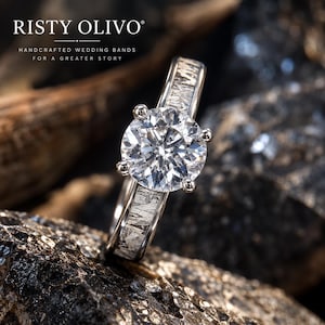 May include: A silver-toned ring featuring a large, round, faceted clear gemstone held by four prongs. The band displays a textured, metallic inlay with a crystalline pattern. The text RISTY OLIVO HANDCRAFTED WEDDING BANDS FOR A GREATER STORY is visible.