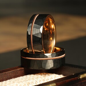 May include: Two black tungsten carbide rings with a rose gold inlay. The rings have a hammered finish and are stacked on top of each other.
