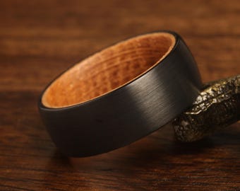 May include: A black ring with a brushed metal finish and a light brown wooden interior. The ring is resting on a textured, bronze-colored surface, set against a dark wood background.