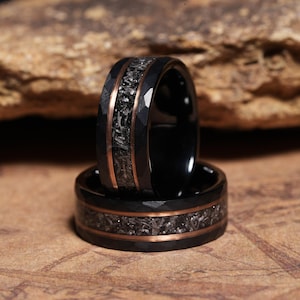 May include: A black ring featuring a hammered finish and a central inlay of dark, textured material bordered by two thin copper-coloured bands. The ring has a smooth interior and a wide, flat profile, showcasing a contrast between the dark metal and the metallic copper accents.