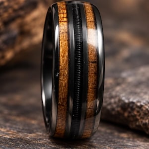 May include: A black metal ring featuring two vertical inlays of brown wood grain and a central thin black grooved band. The polished dark metal edges frame the wood and textured center, creating a contrast between the smooth and ribbed surfaces.
