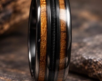May include: A black metal ring featuring two vertical inlays of brown wood grain and a central thin black grooved band. The polished dark metal edges frame the wood and textured center, creating a contrast between the smooth and ribbed surfaces.