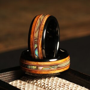 May include: Two black rings with a wooden inlay and abalone shell accents. The rings have a band of copper-colored metal and a polished finish. The rings are displayed on a textured surface.