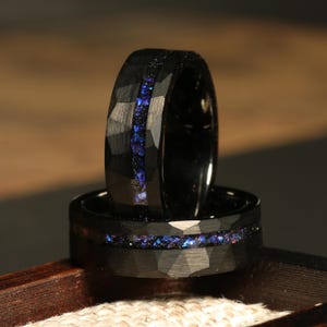 May include: Two black rings with a faceted texture and a central inlay of blue and purple stones. The rings are stacked, showcasing the design and detail of the jewelry. The rings are set against a neutral background.