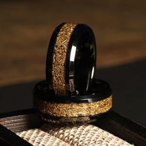May include: Two black rings with a gold-colored textured inlay. The rings are stacked on top of each other, showcasing the intricate design and polished finish. The gold inlay has a rough, organic texture.