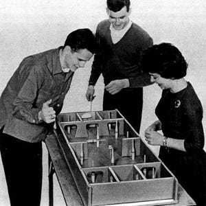 May include: Three people playing a game of tabletop shuffleboard. The game board is made of wood and has a series of numbered lanes. The players are using wooden sticks to push the pucks down the lanes.