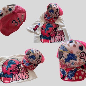 Könnte beinhalten: Eine Sammlung von Artikeln zum Thema Detroit Lions, darunter eine pink-weiße Trucker-Mütze mit der Nummer 313 und ein weißes T-Shirt mit einem pink-blauen Helm-Motiv. Die Mütze und das T-Shirt sind mit Strasssteinen verziert.
