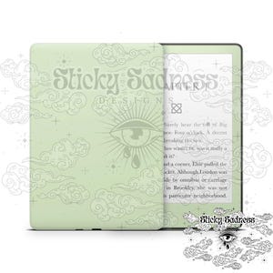 May include: A green Kindle Paperwhite e-reader with a custom case featuring a light green background with a white eye and cloud design. The text "Sticky Sadness Designs" is printed on the case. The e-reader is displaying a page of text from a book.