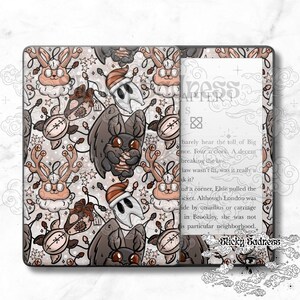 Peut inclure: Une housse de Kindle décorative avec un motif fantaisiste de chauves-souris, de lapins et de personnages de médecins de la peste de dessin animé dans un schéma de couleurs claires. La housse est ouverte, révélant l'écran Kindle avec du texte. La housse a le texte "Sticky Sadness".