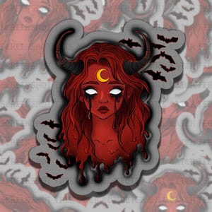 May include: A sticker featuring a red-skinned demon with long red hair, black horns, and a crescent moon on her forehead. The demon has white eyes and black tears, surrounded by black bat silhouettes.