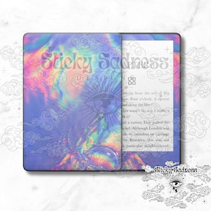 May include: A blue and purple iridescent Kindle Paperwhite e-reader with a holographic sticker featuring a stylized eye and clouds. The text "Sticky Sadness" is visible on the sticker. The e-reader is displaying a page of text from a book.