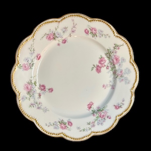 Puede incluir: Un plato de porcelana blanca con borde festoneado y ribete dorado. El plato está decorado con rosas rosas y lavanda y hojas verdes. El plato está sobre un fondo negro.