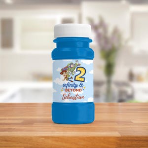 Op de afbeelding: Een blauwe bellenblaasfles met een witte dop. Het etiket toont personages uit een populaire animatiefilm, het nummer 2, de tekst "infinity & beyond" en een naam. De fles staat op een houten oppervlak.